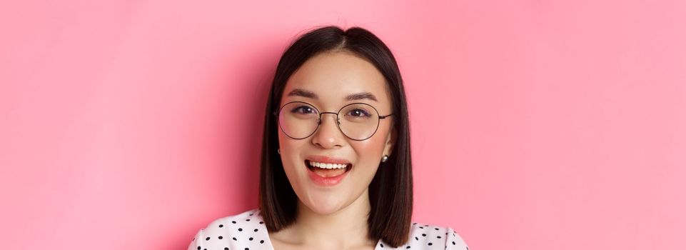 Beauty and lifestyle concept. Close-up of cute asian female model wearing trendy glasses, smiling happy at camera, standing on pink background.