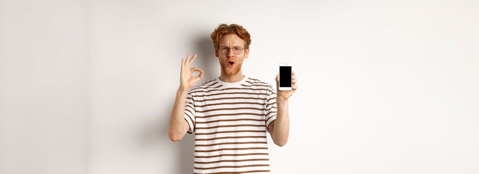 Technology and e-commerce concept. Young man with red hair showing okay sign and blank smartphone screen, praising awesome app or video game, white background.