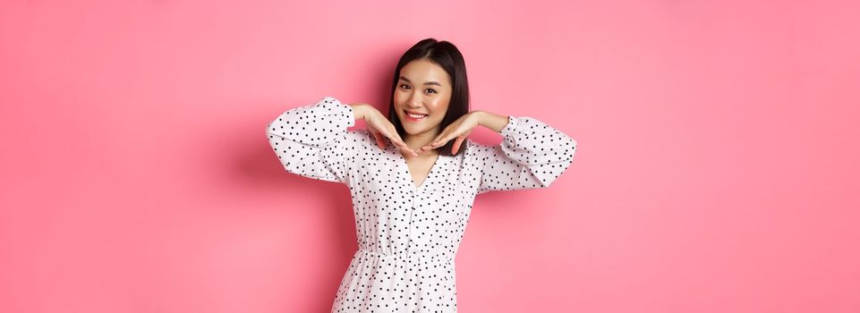 Lovely asian woman showing her beautiful clean face, smiling pretty, standing in dress against pink romantic background.