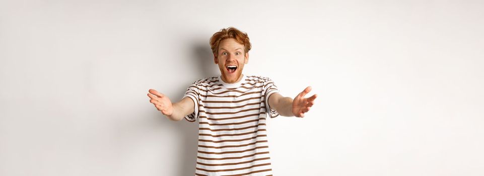 Cheerful man with red curly hair reaching hands forward, stretch out arms to welcome or congratulate you, smiling happy, standing over white background.