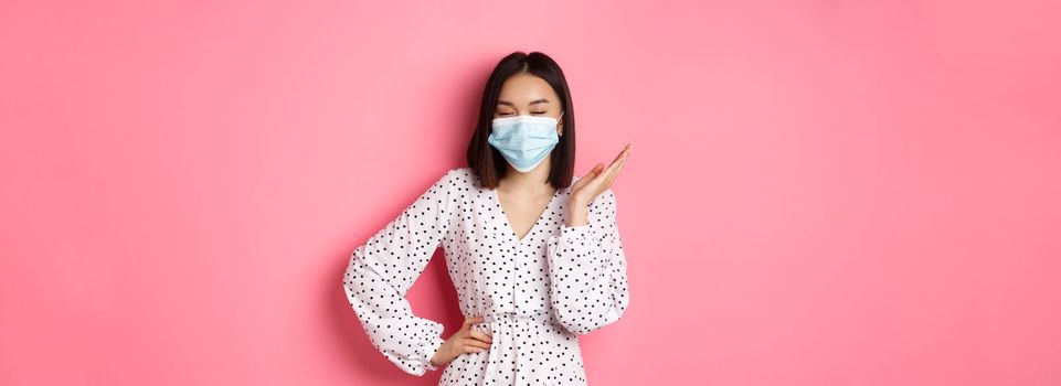 Covid-19, quarantine and lifestyle concept. Beautiful and confident asian woman in dress and face mask looking self-assured, standing sassy against pink background.