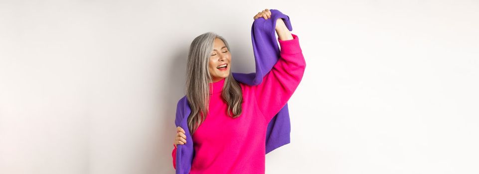 Carefree korean elderly woman in pink sweater, dancing with sweatshirt on shoulders and smiling, posing happy over white background.