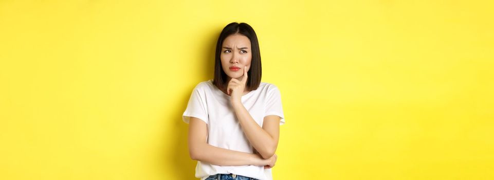 Beauty and fashion concept. Pensive asian woman thinking, looking thoughtful while pondering something, standing over yellow background.