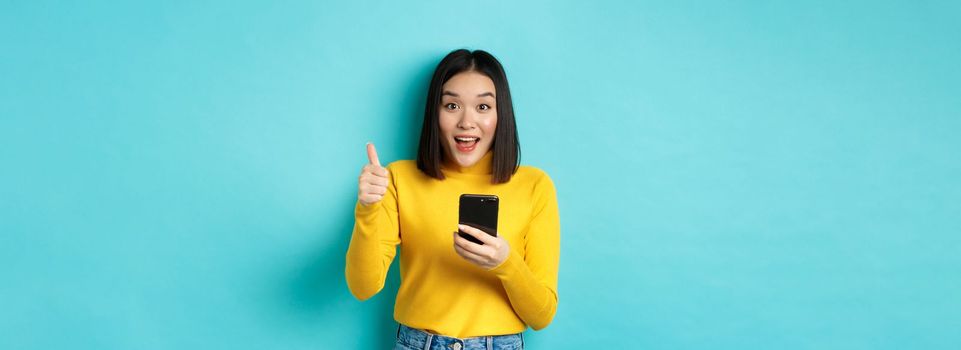 E-commerce and online shopping concept. Excited and amazed asian woman showing thumbs up after using smartphone app, recommend device, standing over blue background.