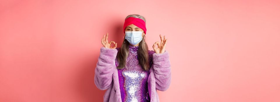 Covid-19, social distancing and fashion concept. Fashionable asian senior woman in face mask and glitter dress, showing okay signs in approval, pink background.