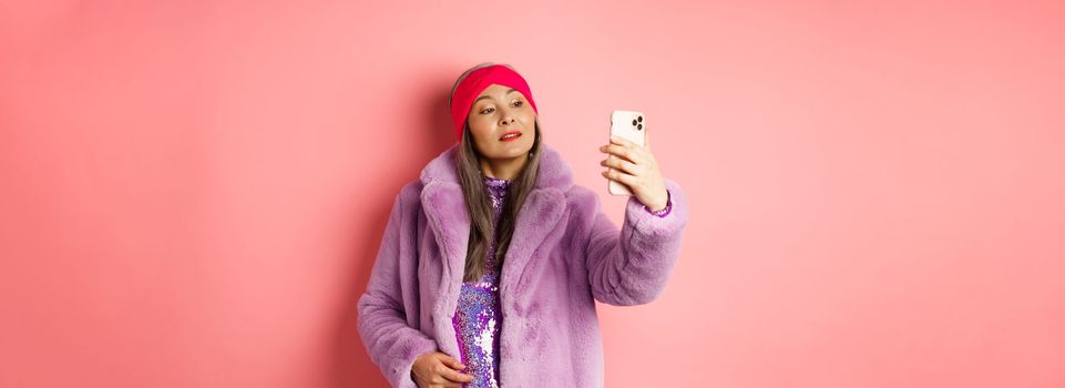 Fashion concept. Stylish asian senior female taking selfie on smartphone, posing in purple faux fur coat and party dress, standing over pink background.