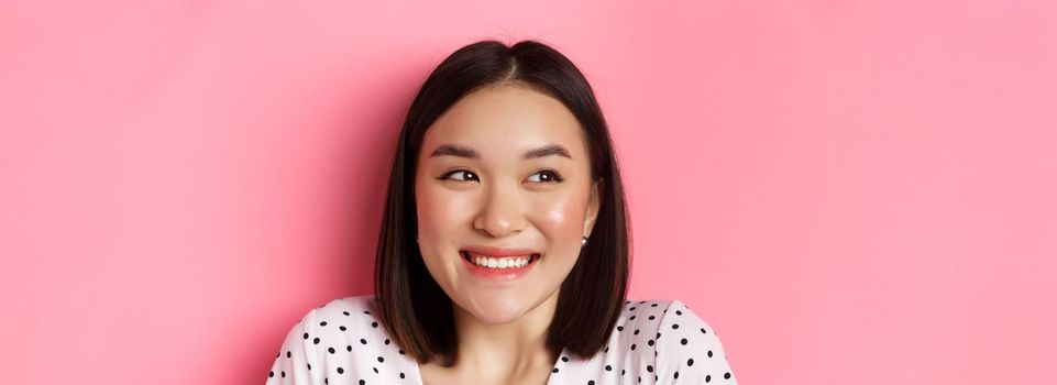 Beauty and lifestyle concept. Headshot of excited asian girl smiling, looking left at promo banner, standing over pink background. Copy space