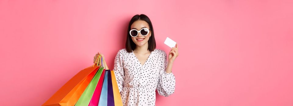 Beautiful asian woman in sunglasses going shopping, holding bags and showing credit card, standing over pink background.