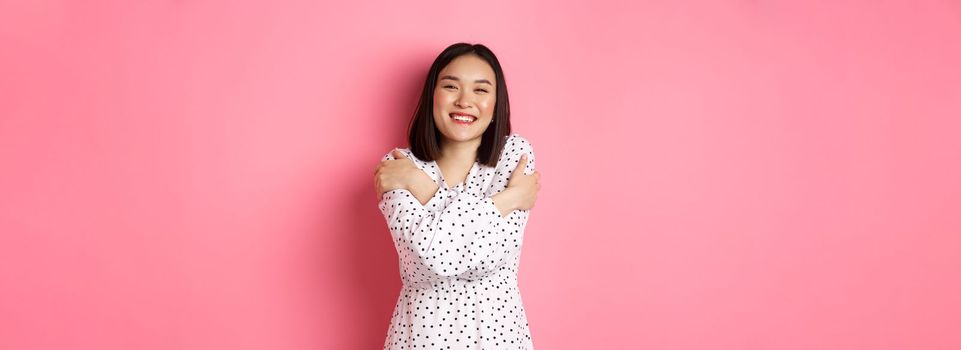 Beautiful asian girl embracing own body, hugging herself and smiling with happy face, looking at camera carefree, standing over pink background.