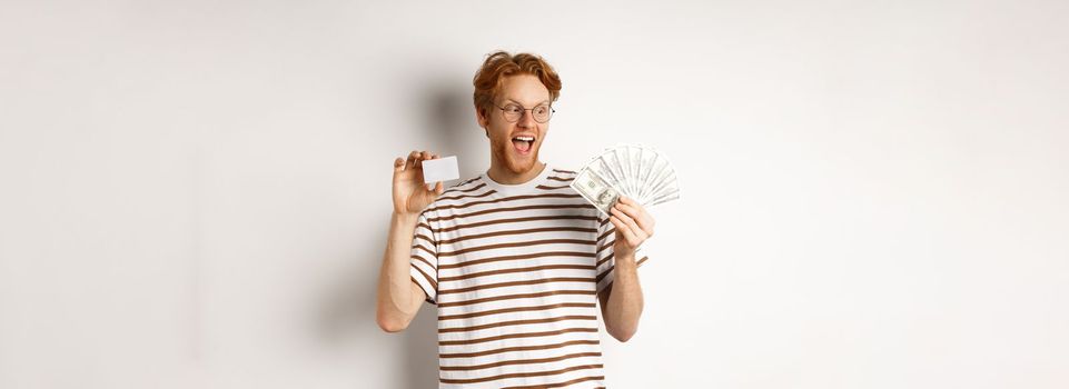 Shopping and finance concept. Cheerful handsome redhead man showing plastic credit card and cash, smiling amazed, white background.