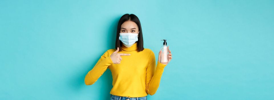 Covid-19, social distancing and pandemic concept. Cheerful asian woman showing hand sanitizer, recommend use antiseptic from coronavirus, wearing medical mask.