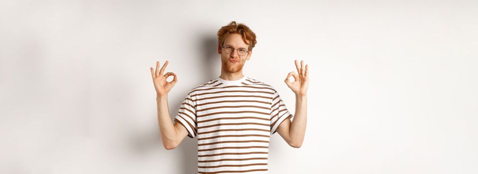 Attractive redhead guy in glasses showing okay signs and looking impressed, praising something cool, standing over white background.