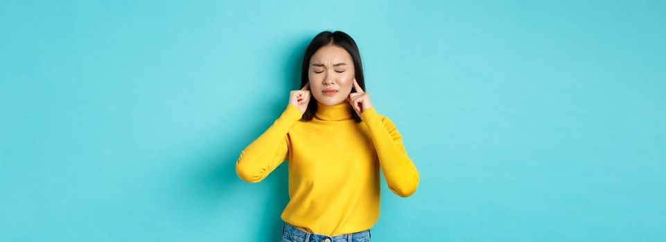 Tired and disappointed young asian woman unwilling to listen, shut ears with fingers and close eyes, standing in denial over blue background.