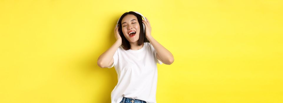 Attractive asian woman enjoying listening music in wireless headphones, smiling pleased and singing along, yellow background.