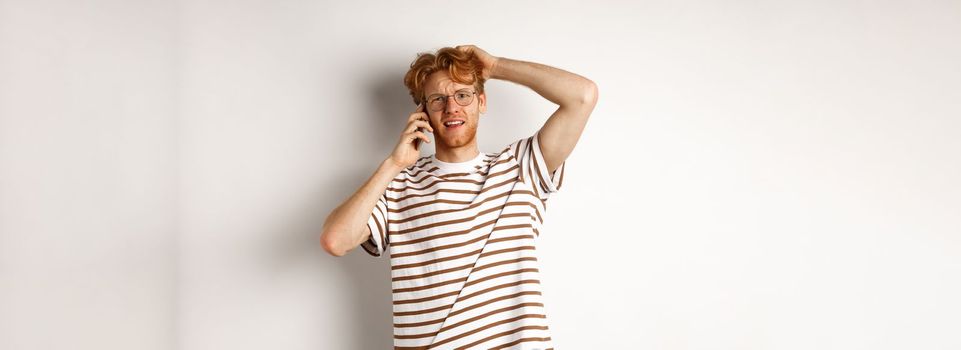 Puzzled redhead guy talking on phone, scratching head and looking confused or indecisive, standing over white background.