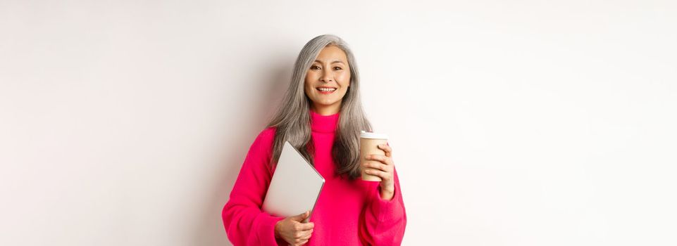 E-commerce concept. Happy asian senior freelancer, drinking coffee and holding laptop, smiling relaxed at camera, standing over white background.