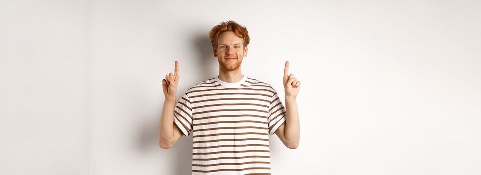 Smiling redhead guy with beard, pointing fingers up and showing advertisement, standing over white background.