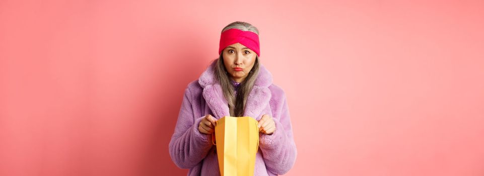 Upset senior asian woman open shopping bag and looking sad at camera, standing over pink background.