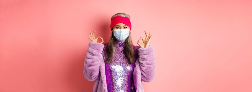 Covid-19, social distancing and fashion concept. Fashionable asian senior woman in face mask and glitter dress, showing okay signs in approval, pink background.