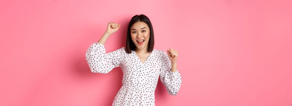 Beautiful korean woman dancing and having fun, smiling happy at camera, posing against pink background.