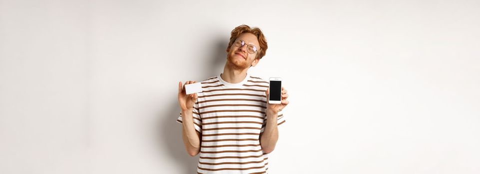 Shopping and finance concept. Pleased young man with red hair smiling from satisfaction, showing smartphone blank screen and credit card, white background.