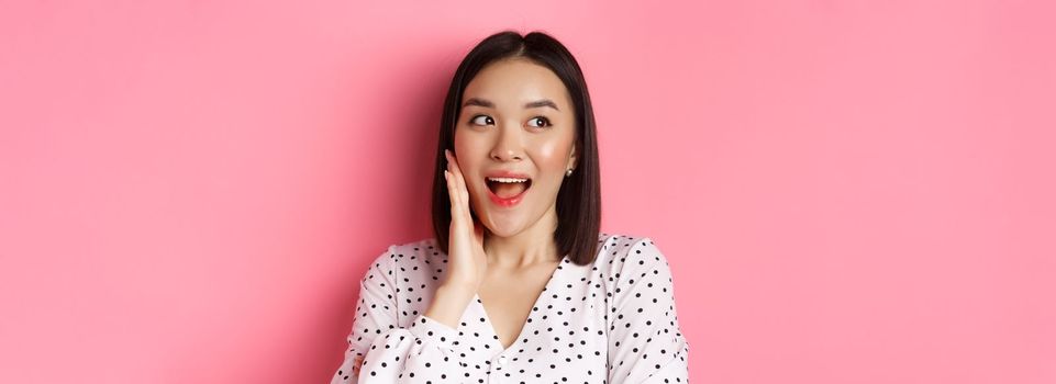 Coquettish asian woman in blushing, touching cheek and looking left amused, standing over pink background.