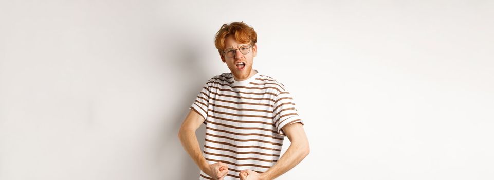 Image of confident and strong redhead man flexing biceps, showing muscles after gym, standing over white background.