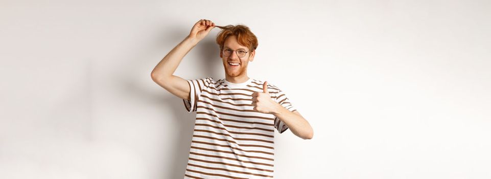 Cheerful redhead guy showing thumb-up and his red hair strand, like new color, standing over white background.