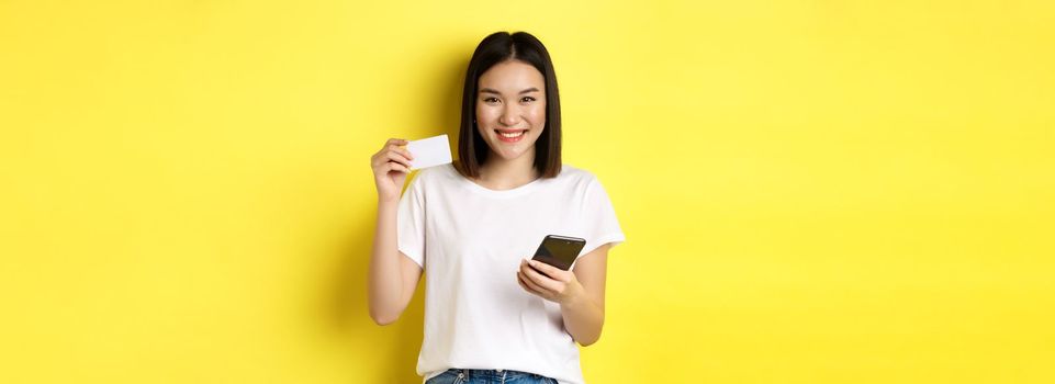 Attractive korean woman paying online with smartphone, showing plastic credit card and smiling, standing over yellow background.