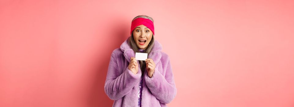 Shopping and fashion concept. Beautiful asian middle-aged woman showing plastic credit card and smiling amazed in stylish clothes, pink background.