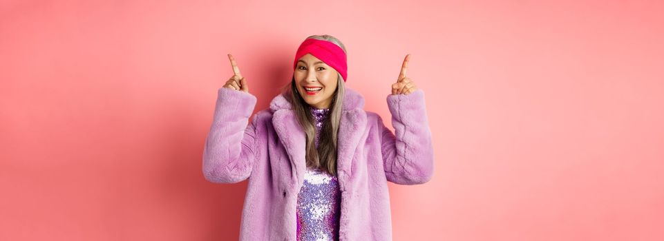 Stylish asian woman smiling happy, pointing fingers up and showing advertisement, wearing winter fake-fur coat and headband, pink background.