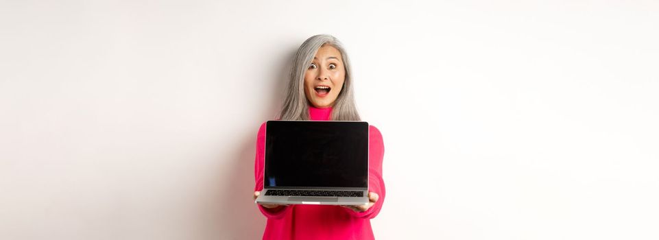 E-commerce concept. Amazed asian senior woman showing blank laptop screen and looking happy, demonstrating promo, white background.