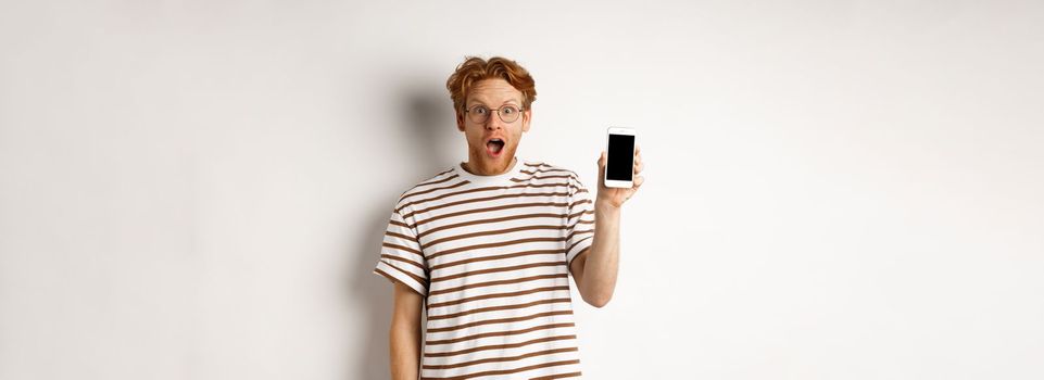 Technology and e-commerce concept. Surprised and shocked redhead guy checking out online promotion, showing blank smartphone screen and drop jaw, white background.