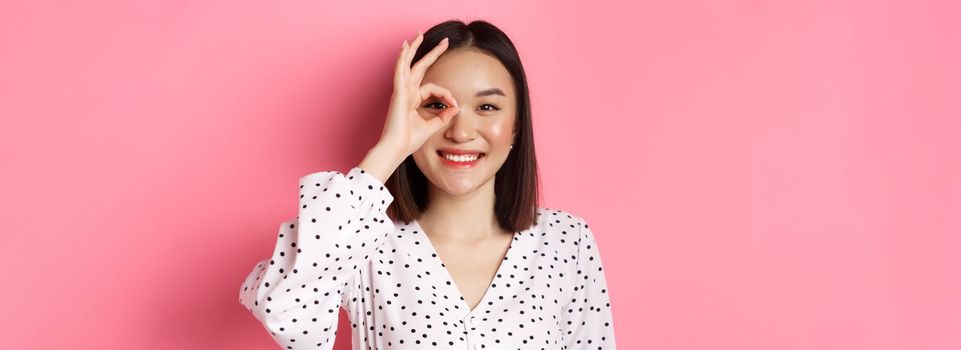 Beauty and lifestyle concept. Close-up of cute smiling asian girl showing okay sign on eye, standing over pink background in dress.