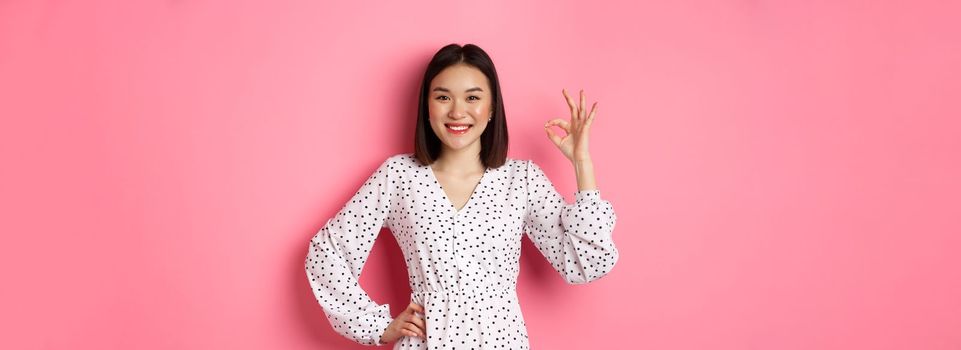 Pretty young asian woman in dress showing okay sign, praising and showing approval, looking satisfied, standing against pink background.
