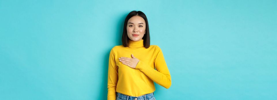 Image of proud smiling asian woman holding hand on heart, showing respect to national anthem, standing over blue background.