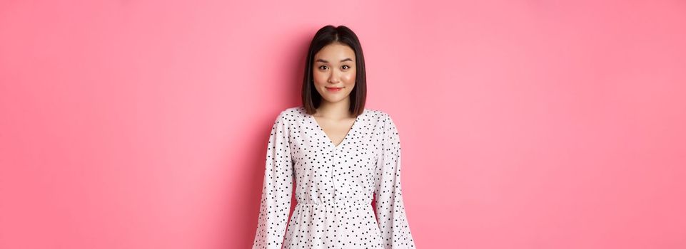 Beautiful asian lady smiling at camera, wearing cute romantic dress, standing over pink background. Copy space