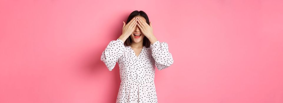 Happy romantic asian girl waiting for surprise, close eyes and smiling amazed, anticipating something, standing over pink background.