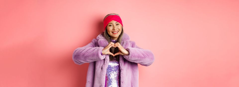 Romance and valentines day. Happy asian senior woman showing heart sign, I love you gesture, smiling and looking caring at camera, standing in faux fur coat, pink background.