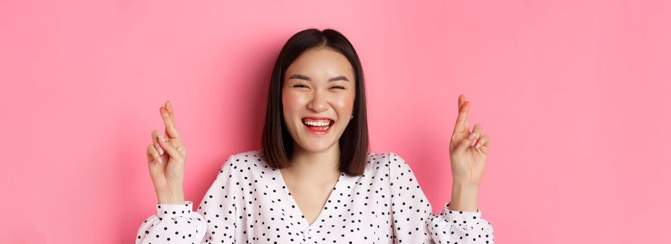 Beauty and lifestyle concept. Close-up of happy asian woman making a wish, holding fingers crossed for good luck and smiling optimistic, standing over pink background.