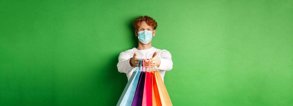 Happy young man in medical mask giving you shopping bags with purchases, smiling and wishing well, standing over green background. Covid-19 concept.