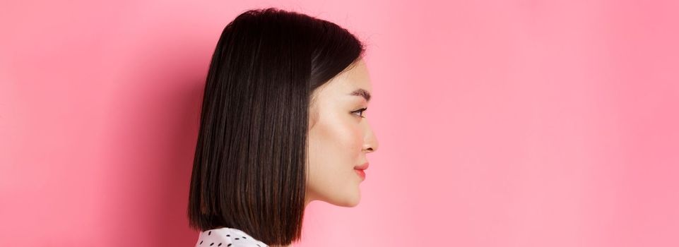 Beauty and skin care concept. Headshot profile of young beautiful asian woman with short dark hair, looking left at copy space, standing over pink background.