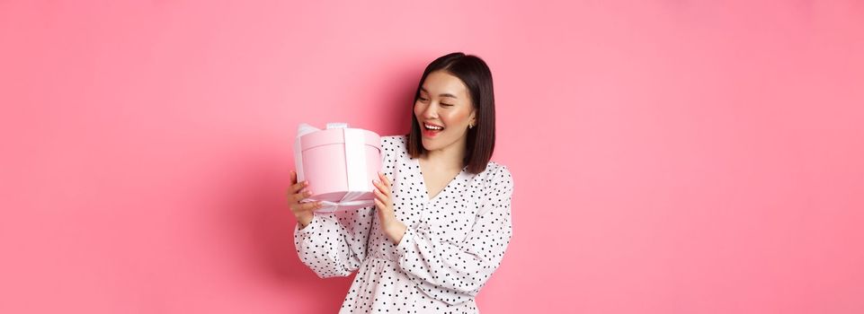 Valentines day, celebration concept. Beautiful asian woman holding romantic gift box, smiling happy, standing over pink background.