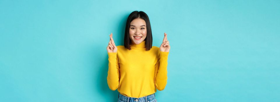 Hopeful and optimistic asian woman cross fingers for good luck, making wish and smiling, looking at camera and expecting something, standing over blue background.