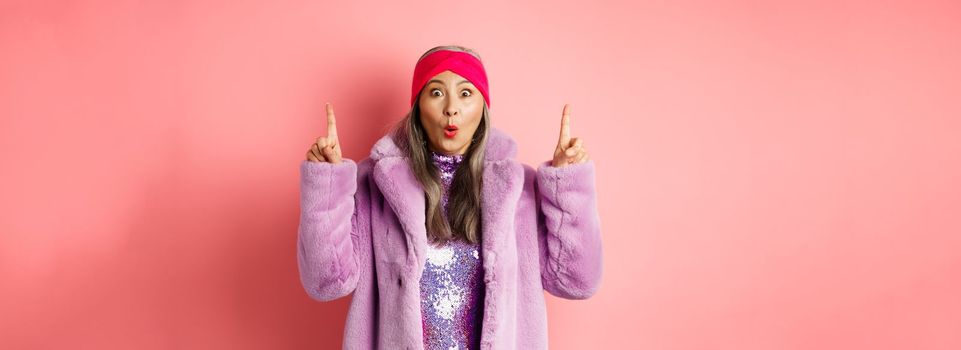 Wow check this out. Amazed asian senior woman pointing fingers up, staring at camera impressed, showing fashion or beauty advertisement, pink background.