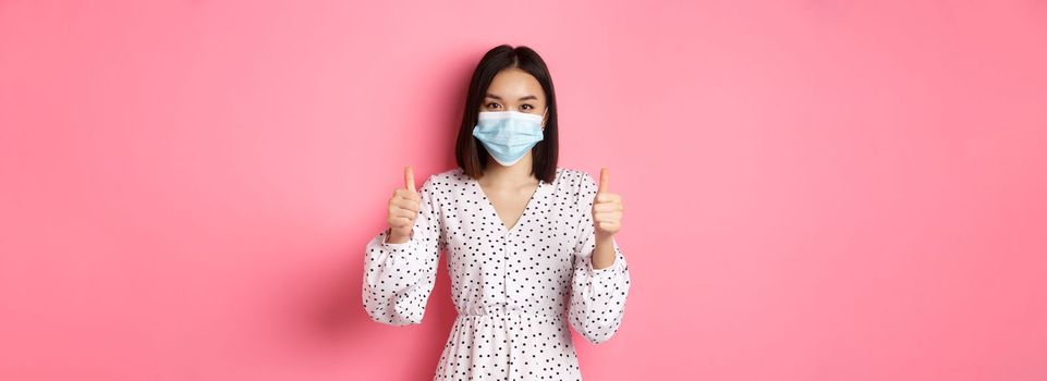 Covid-19, quarantine and lifestyle concept. Beautiful asian woman in dress and face mask showing thumbs-up, give approval, like and praise, standing over pink background.