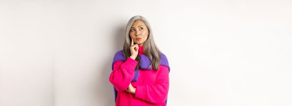 Thoughtful asian middle-aged mother looking upper left corner, thinking with serious face, standing over white background. Copy space