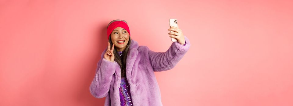 Fashion concept. Cool and cheerful asian senior woman taking selfie with peace sign, wearing trendy faux fur coat and party dress, standing over pink background.