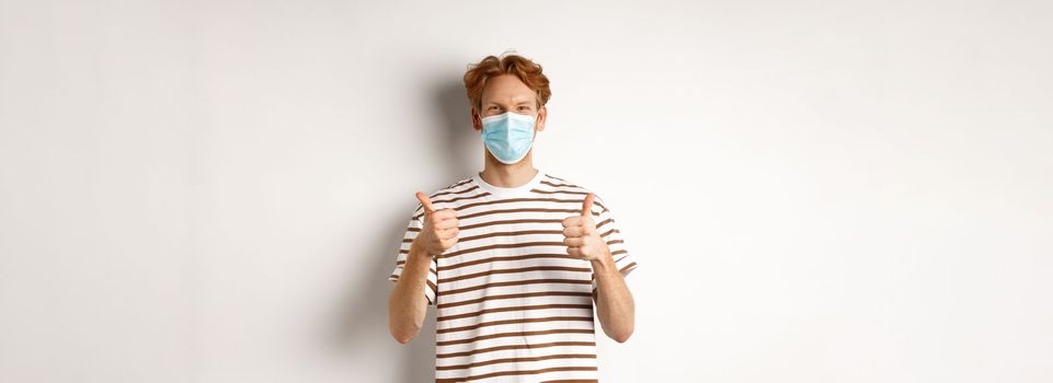 Covid-19, pandemic and social distancing concept. Young man with red hair wearing medical mask to prevent catching coronavirus, showing thumbs-up, white background.