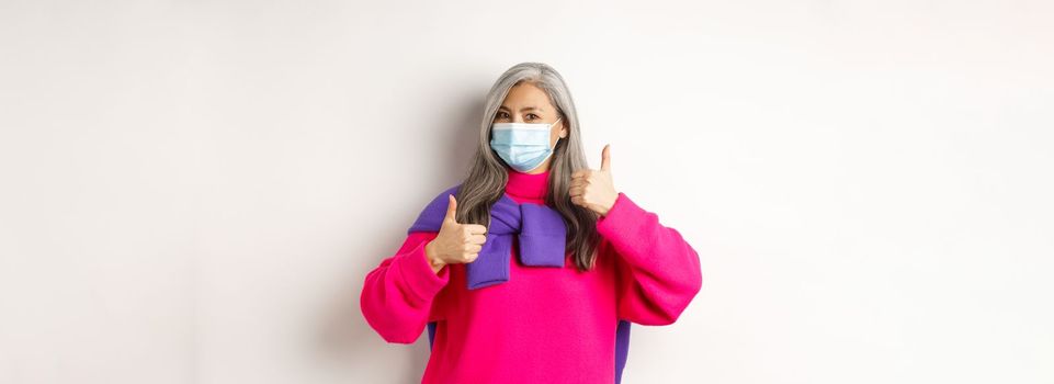 Covid, pandemic and social distancing concept. Cheerful and stylish asian senior woman wearing medical mask and showing thumbs-up, standing over white background.